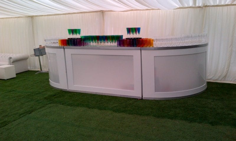 Interior of a traditional marquee with a large white oval illuminated bar counter and rows of colourful cocktail glasses