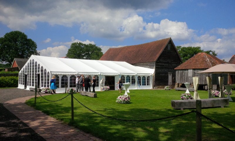 A clearspan marquee beside an old English barn and granary at a sunny wedding reception — marquee hire in Kent
