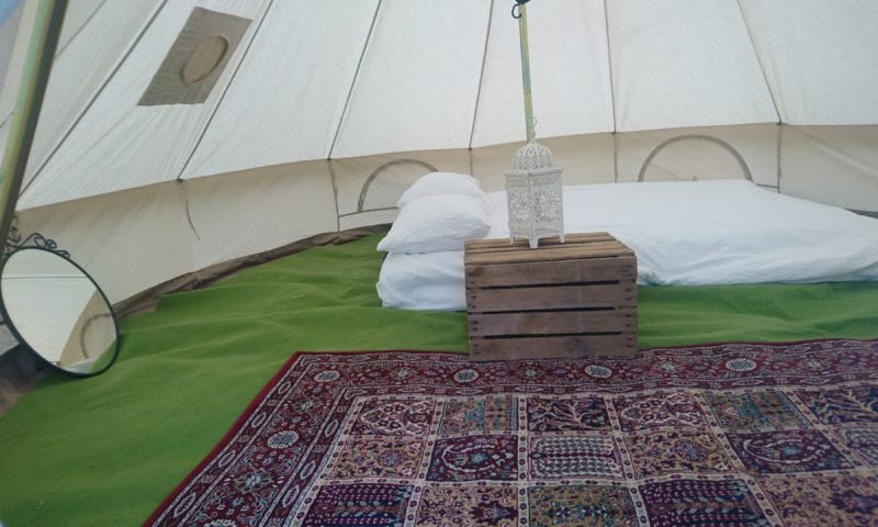 Bell tent interior set up as a glamping bedroom — Persian rug, pallet bedside table, Moroccan lantern and white pillows on a low mattress