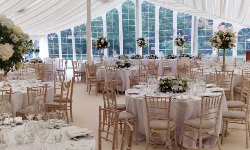 Elegant clearspan marquee wedding breakfast from a wide angle — Gothic arched windows, pale linen and gold candelabra