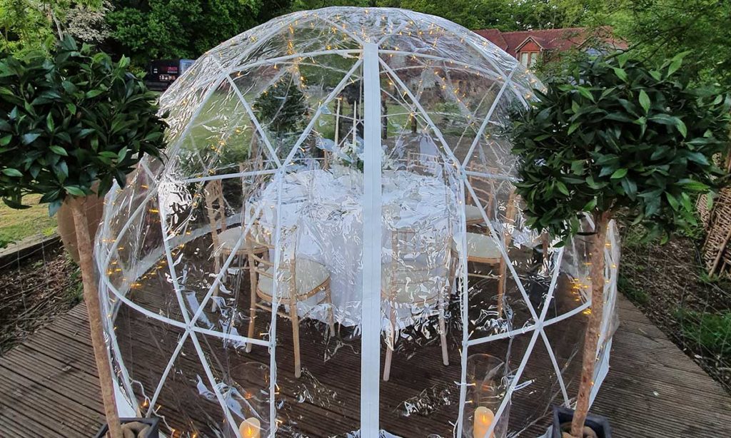 Aerial view of a clear geodesic dome on a timber deck with topiary bay trees flanking the entrance