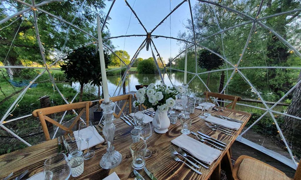 Interior of a clear dome in a sunny English garden with a river view — rustic trestle table and white ranunculus flowers