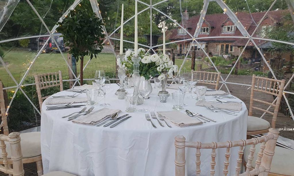 Interior of a clear geodesic dome at dusk — a round table for eight with white linen and crystal glassware