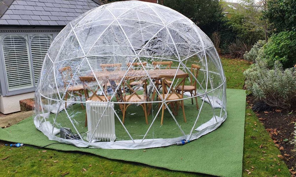 Exterior of a clear dome on artificial grass — large rustic dining table visible inside, period house with conservatory behind