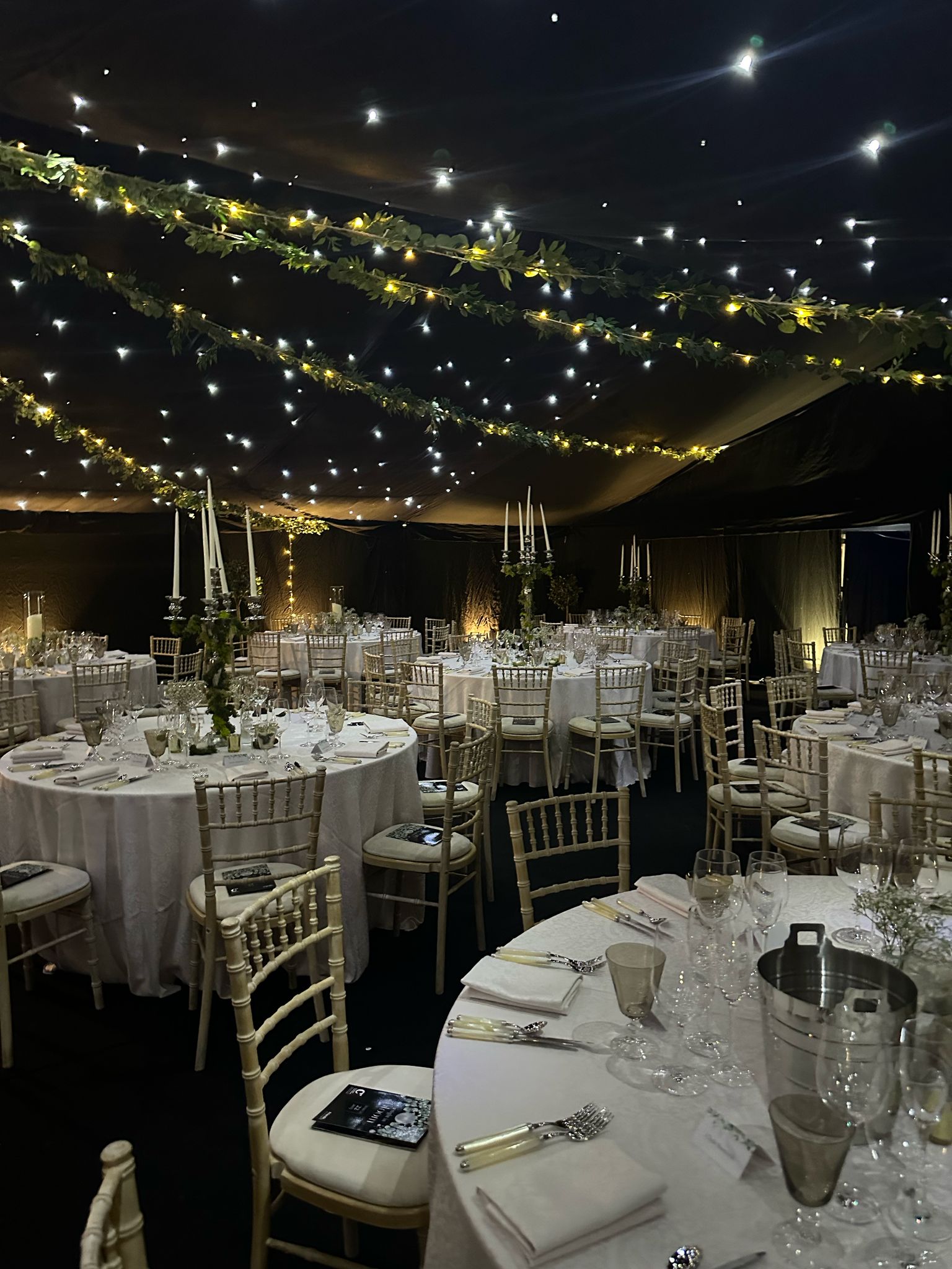 Formal dining marquee at night with round tables, Chiavari chairs, starry ceiling lights and hanging greenery