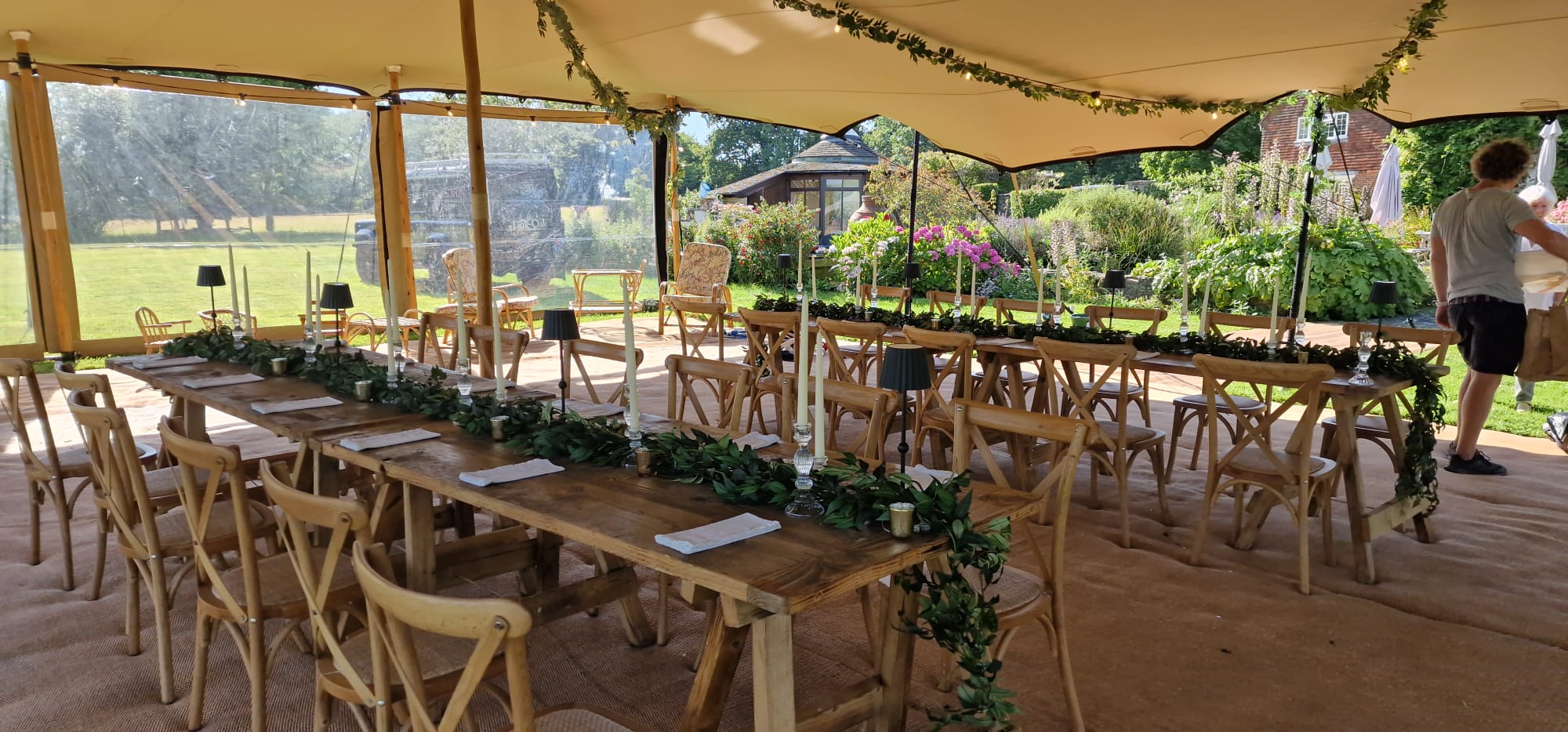 Open-sided marquee dining setup with trestle tables, wooden chairs and greenery table decor