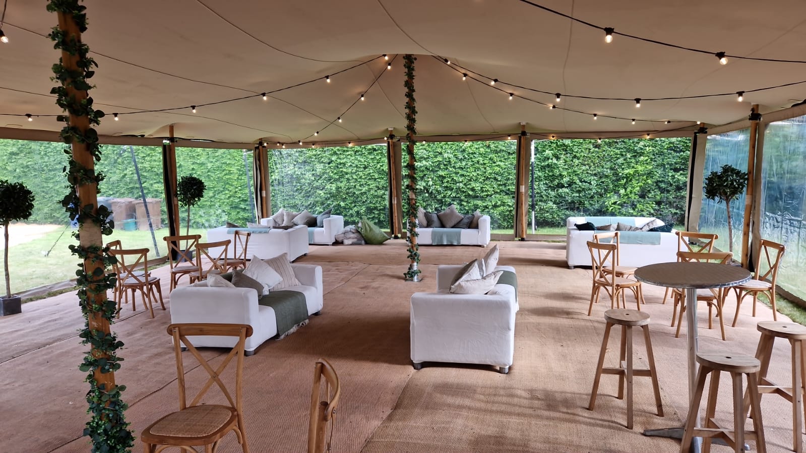 Open-sided marquee interior with sofas, poseur tables, festoon lights and greenery-wrapped supports