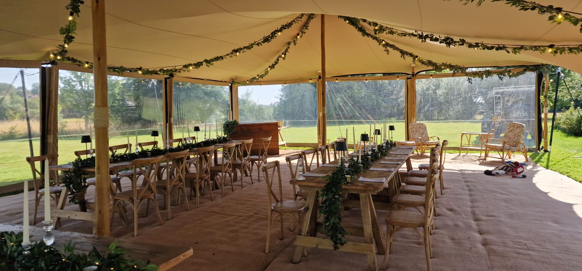 Open-sided marquee with long trestle tables, wooden chairs, greenery garlands and views over the lawn