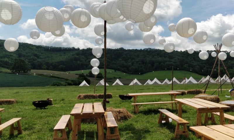 Large white paper lanterns strung over a hillside field with bell tents and tipis in the pastoral distance