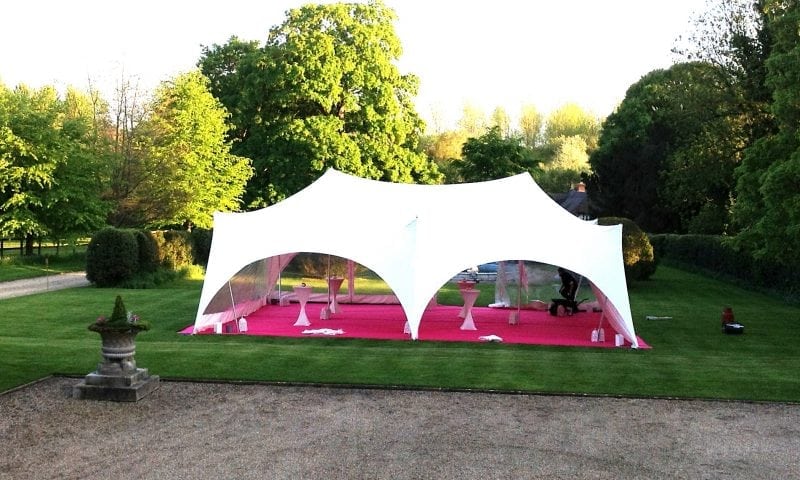 A pagoda marquee at dusk with vivid pink and fuchsia uplighting glowing through its fabric walls in a formal garden