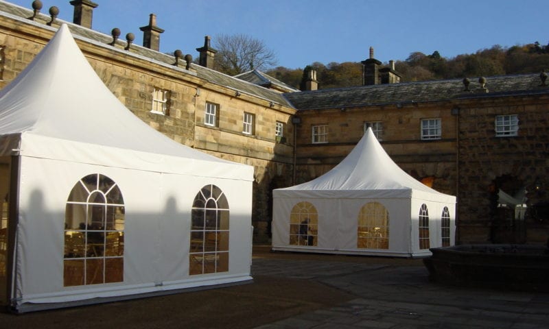 Two white pagoda marquees with enclosed sides and Georgian arched windows in the stone courtyard of a grand country house