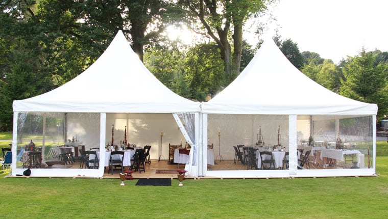 A double linked pagoda marquee with clear sides in an English country garden at late afternoon, dining tables visible within