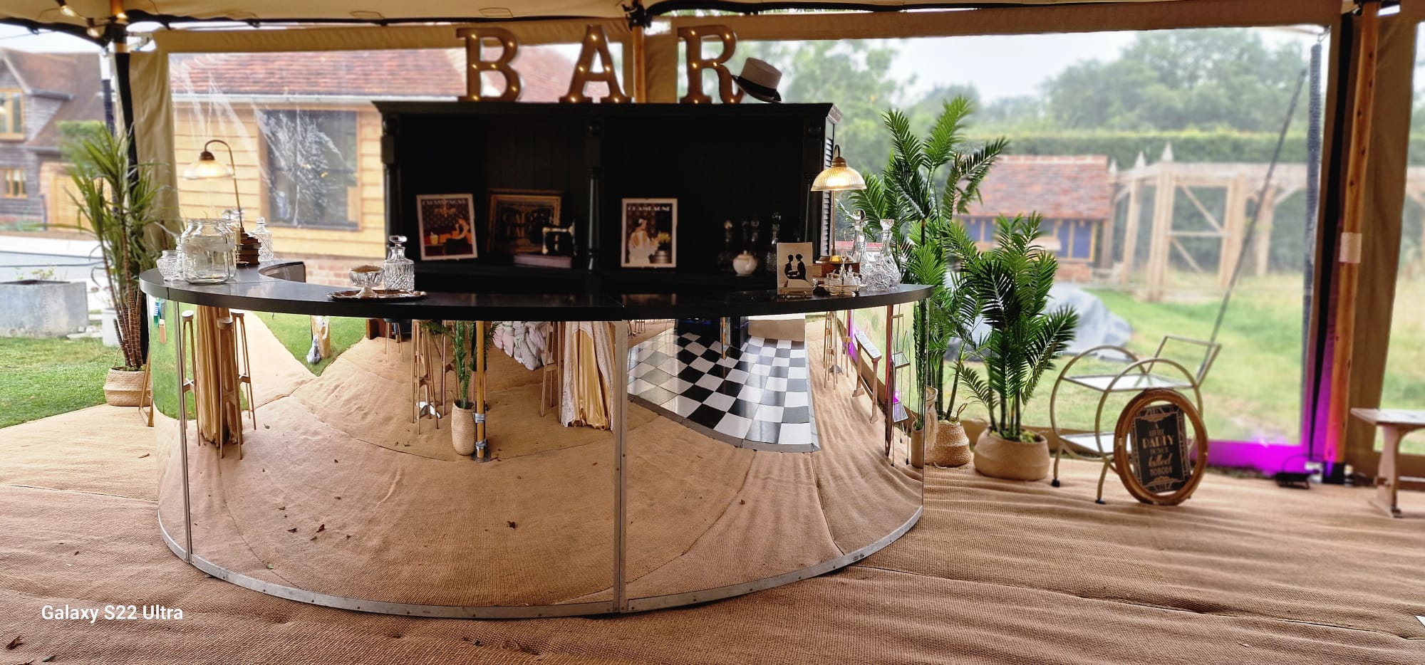 Bar area inside a rustic marquee with a mirrored bar front, plants and framed decor behind
