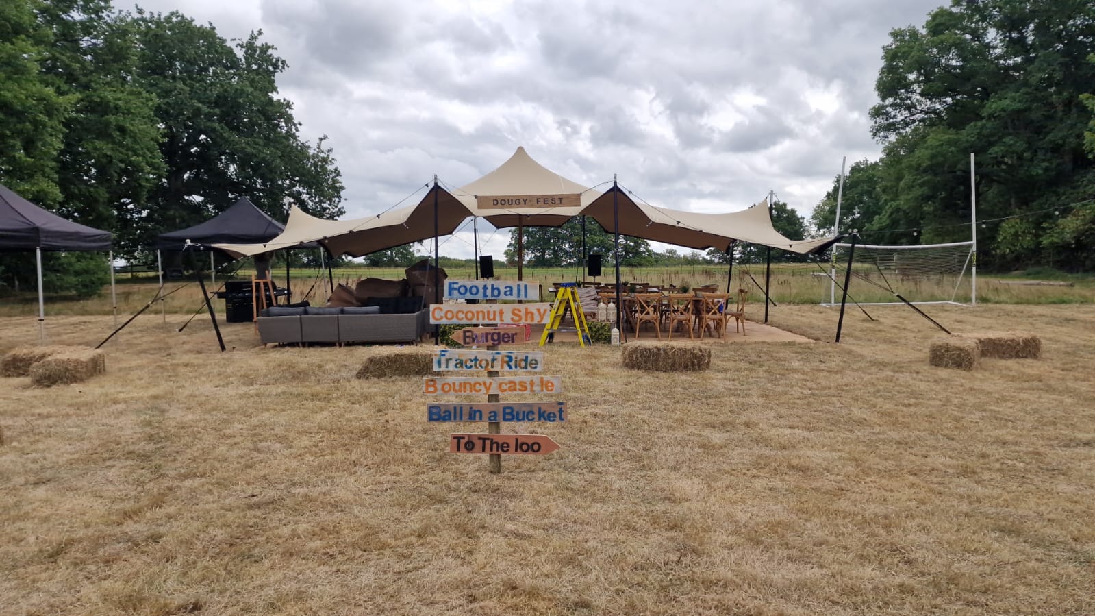 Rustic outdoor event setup with multiple canopy peaks, bunting-style signage and seating on dry grass