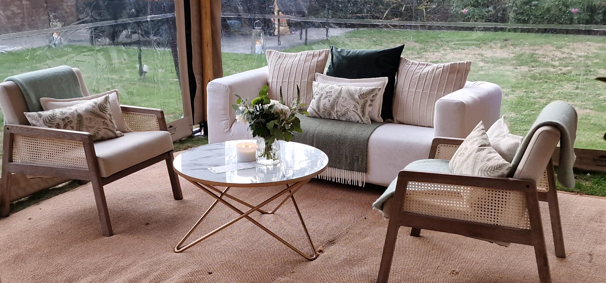 Cosy lounge corner inside a stretch tent — a cream sofa with patterned cushions, cane armchairs, a marble coffee table and jute rug, garden visible through clear walls