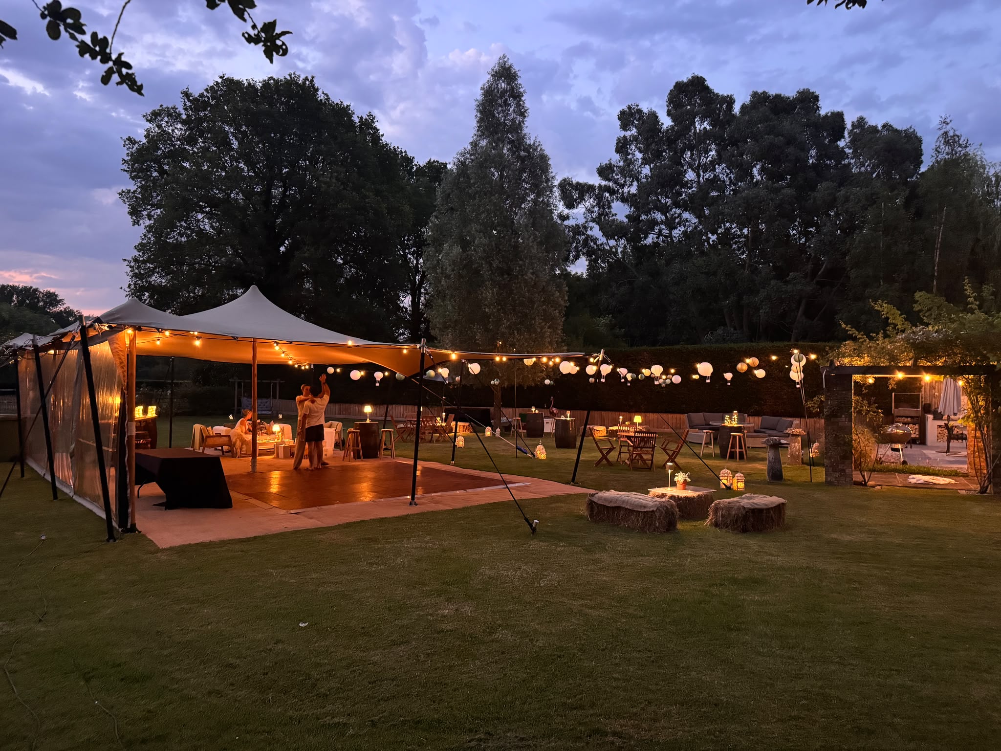 An open-sided stretch tent at dusk with a dance floor, round paper lanterns and festoon lights, tree-stump stools arranged on the lawn outside