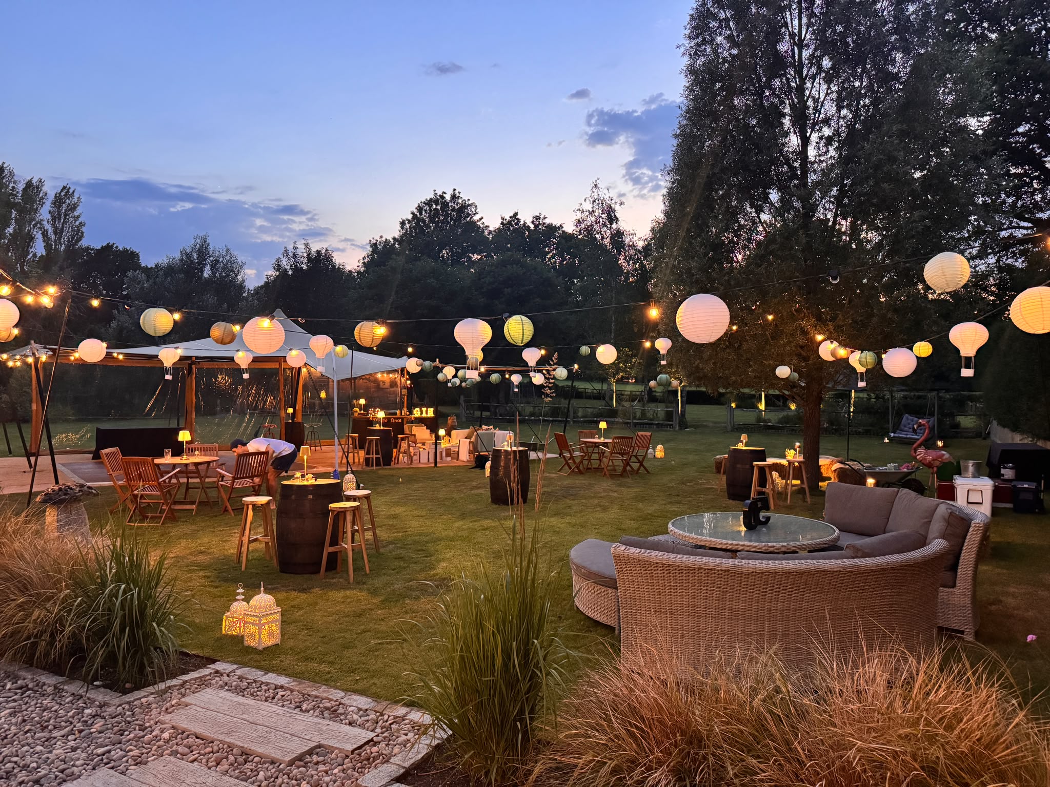 A garden at dusk beside a stretch tent — paper lanterns and festoon lights glowing over a rattan daybed, oak-barrel tables and poseur stools