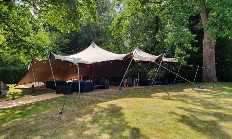 Exterior of a stretch tent in a wooded English garden with festoon lights strung along the support cables