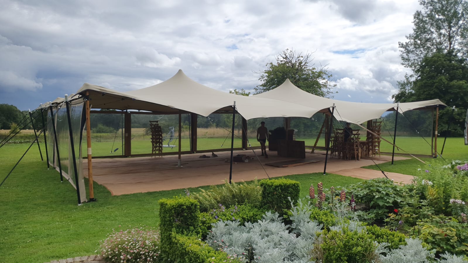 A cream stretch tent on a manicured lawn beside a herbaceous garden border, open-sided with timber poles and stacked chairs ready for an event