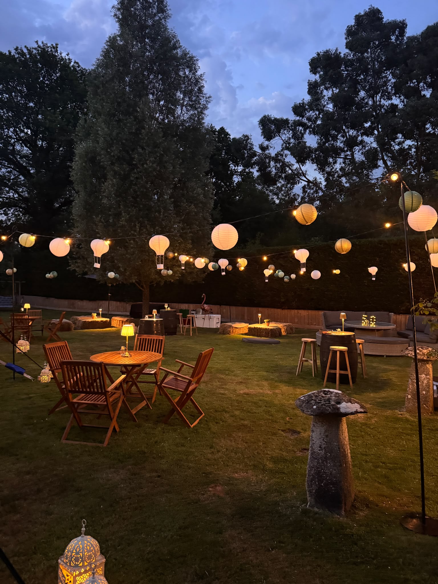 An evening garden party with round timber tables and chairs beneath a canopy of paper lanterns and festoon lights