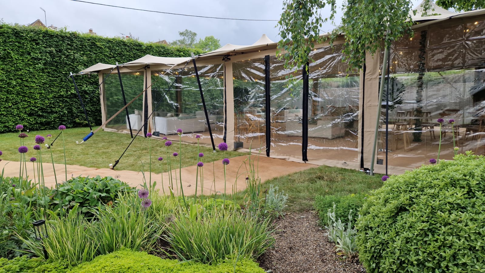 Stretch-style garden marquee beside landscaped planting, with clear walls and timber poles over a patio area
