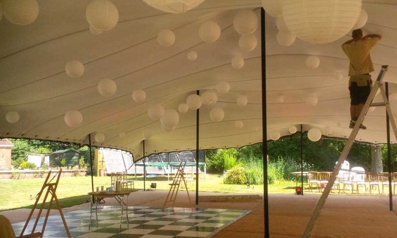 A worker on a ladder inside a stretch tent hanging white paper lanterns while a chequered dance floor is being laid below