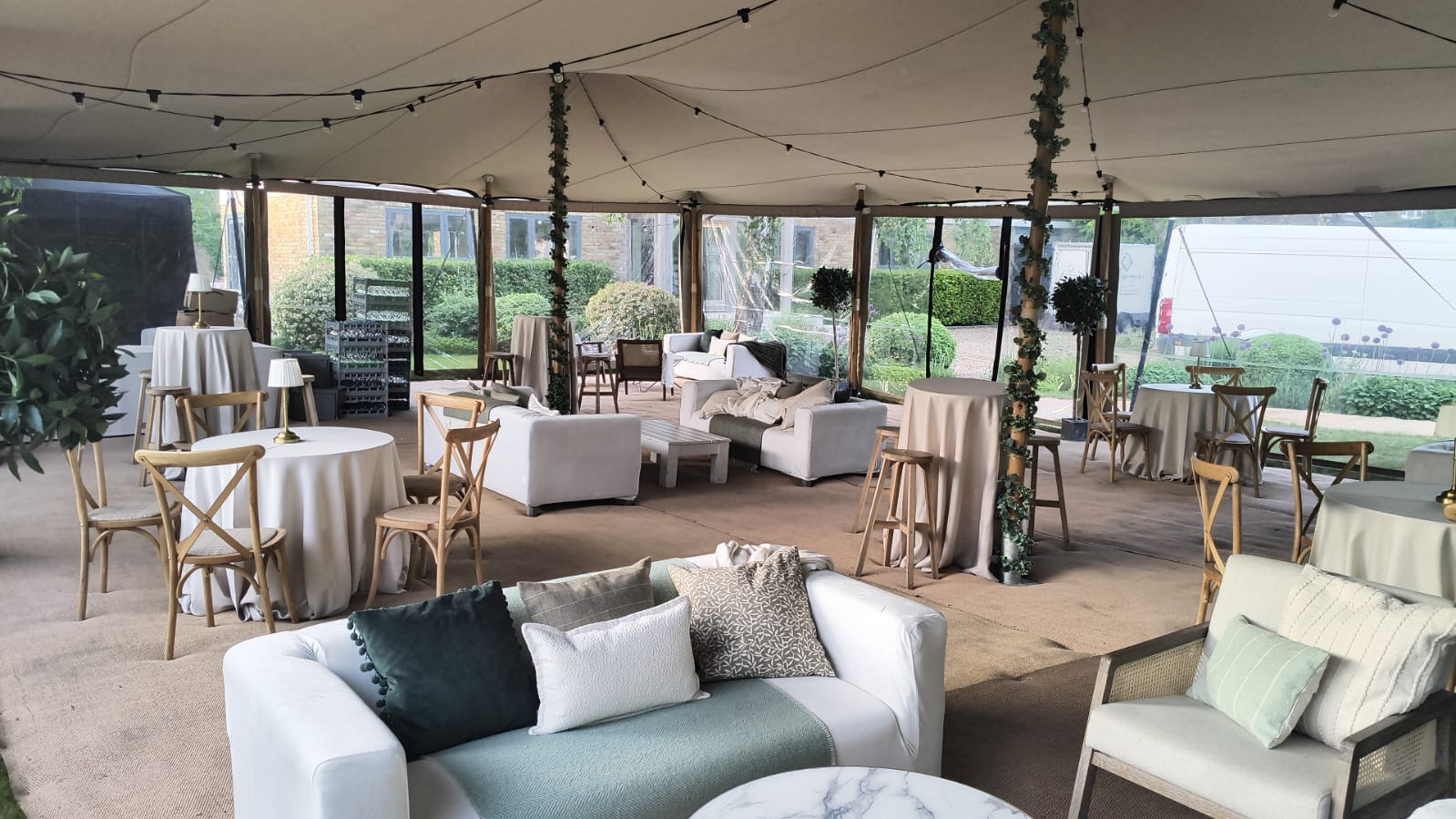 Stretch tent lounge interior with cream sofas, poseur tables, cross-back stools and ivy-wrapped support poles, clear walls looking onto a garden