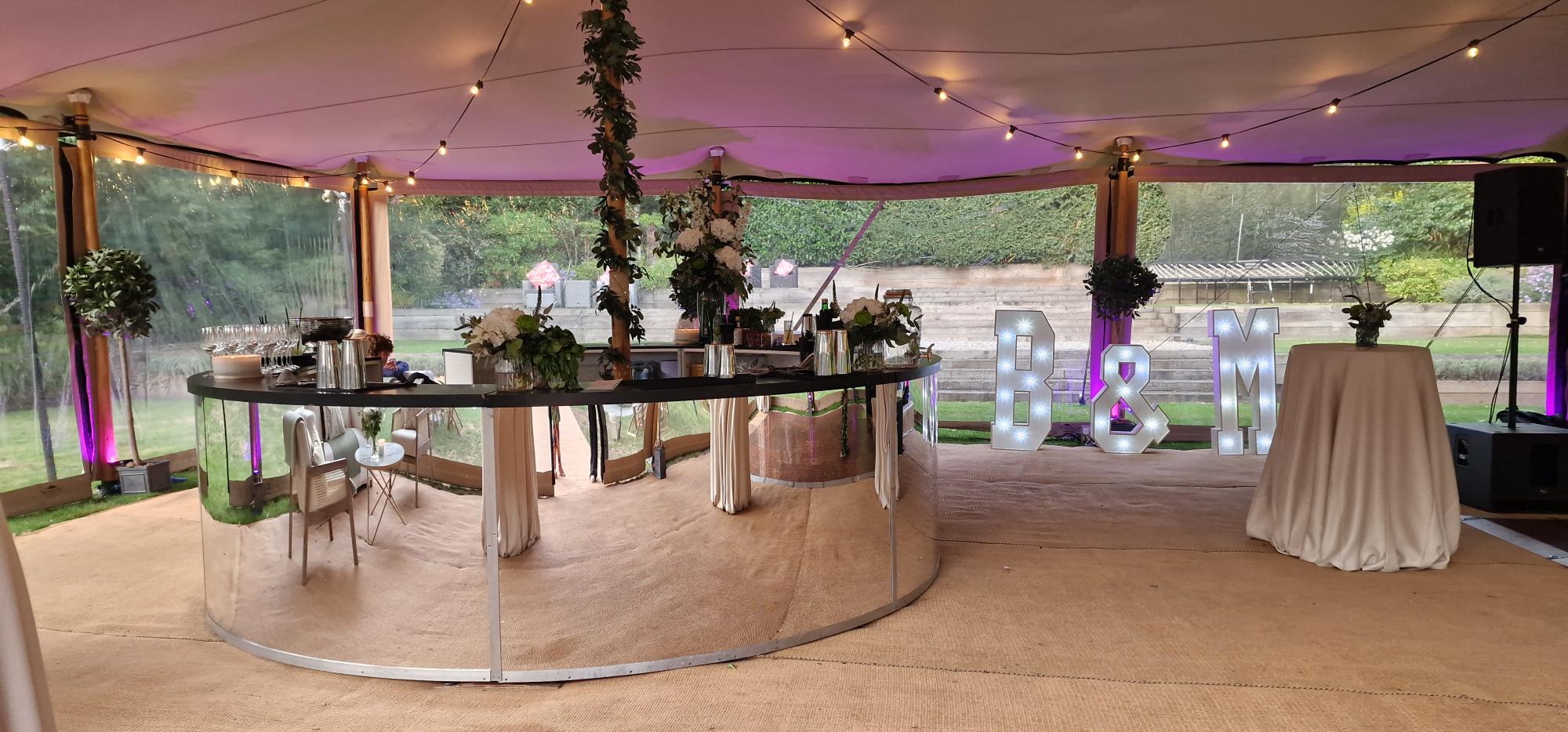 Evening inside a stretch tent — a curved mirror-fronted bar wrapped in greenery, festoon lights overhead and illuminated B&M letters on the dance floor