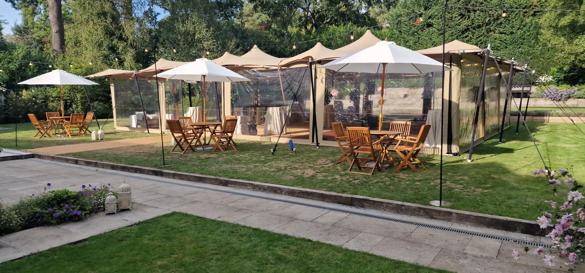 A run of open-sided canopy peaks and parasols on a lawn with timber dining tables and chairs in dappled afternoon light