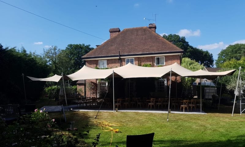 A stretch tent covering a patio with cross-back chairs set for dining, English country house behind