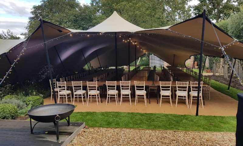 Open-sided stretch tent in a walled garden with chiavari chairs, dining tables and a cast iron fire pit outside