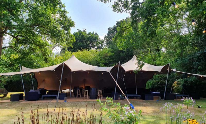 Exterior of a dark khaki stretch tent in an English wooded garden between tall trees, open-sided with dark lounge furniture inside