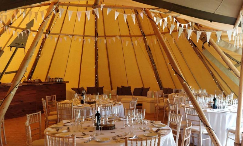 Interior of a tipi with pale bunting strung between the poles, round tables with white linen and natural wood chiavari chairs