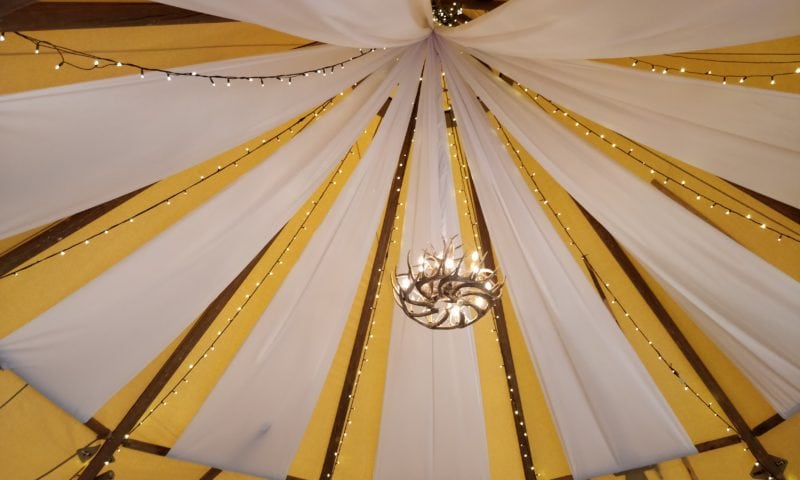 Looking up inside a tipi — alternating white fabric and golden canvas panels with fairy lights along each rib