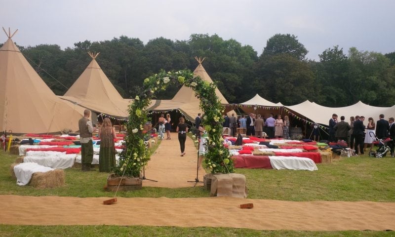 Wedding guests arriving at a tipi complex through a floral arch with hay bales, bunting and a grassy field behind