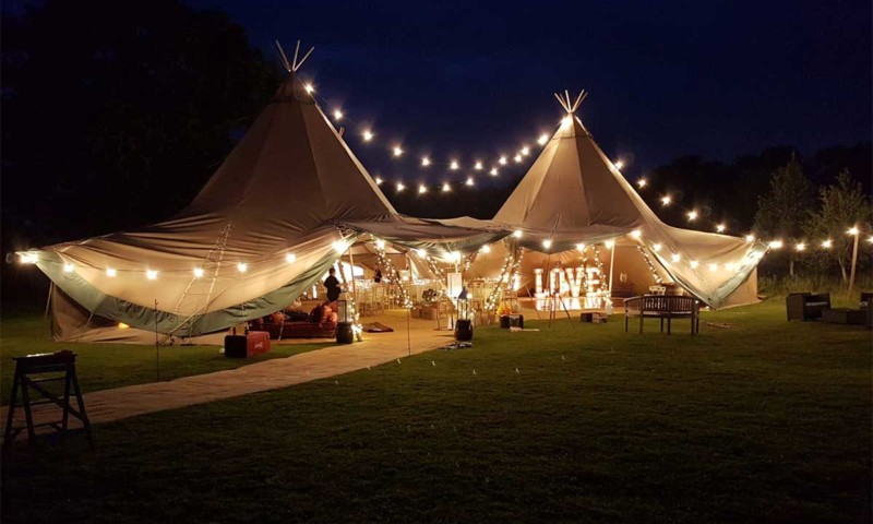 Exterior of a double tipi at night with festoon and Edison lights strung outside — an illuminated LOVE sign at the entrance