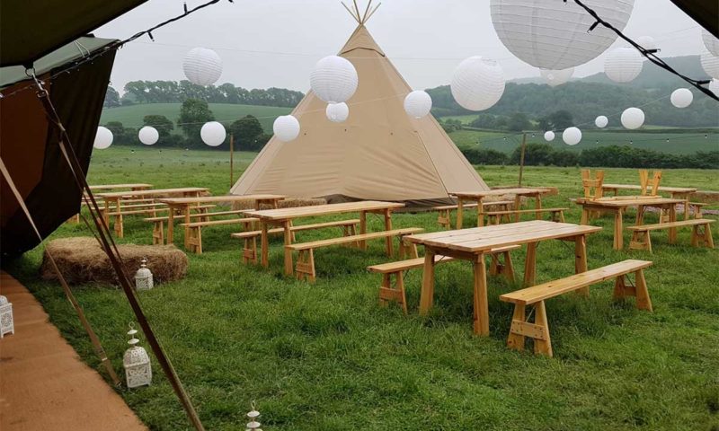 View from inside a tipi toward a meadow, with hardwood trestle tables and benches set up for dining