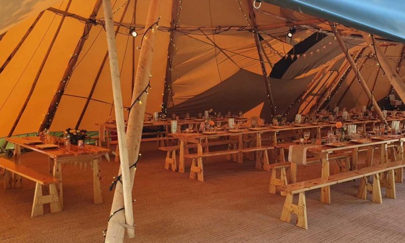 Inside a tipi at night — long hardwood trestle tables with benches running the full length, fairy lights along the poles