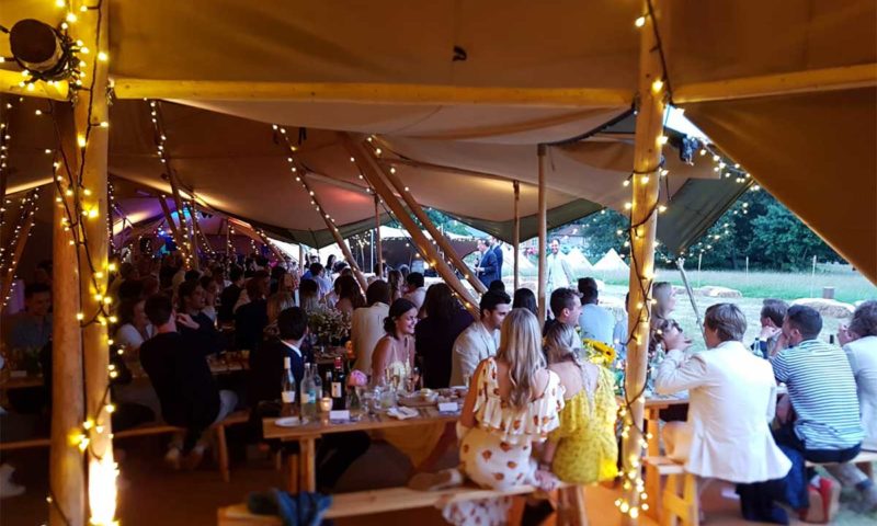 A large linked tipi filled with wedding guests at long trestle tables with fairy lights
