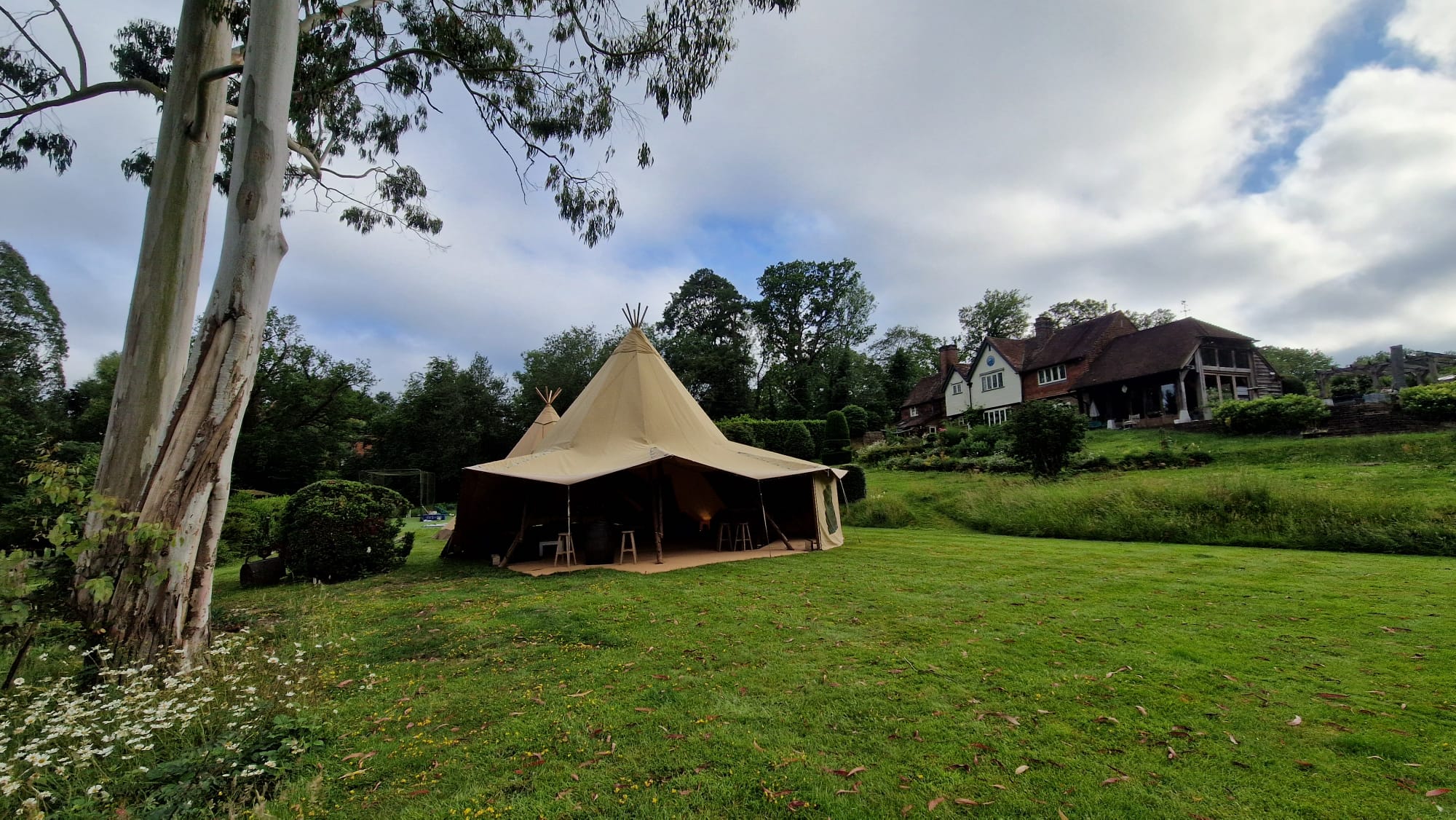 A single tipi pitched on a sloping lawn with a large tile-hung country house in the background on an overcast day