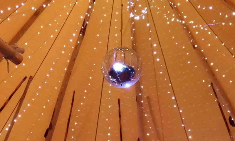 Looking up inside a tipi — golden canvas ceiling, fairy lights along the structural ribs and a mirror ball at the peak