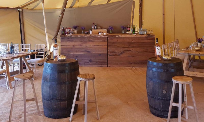 Inside a tipi — two oak barrel poseur tables with pine stools and a dark wood bar stocked with bottles and flowers
