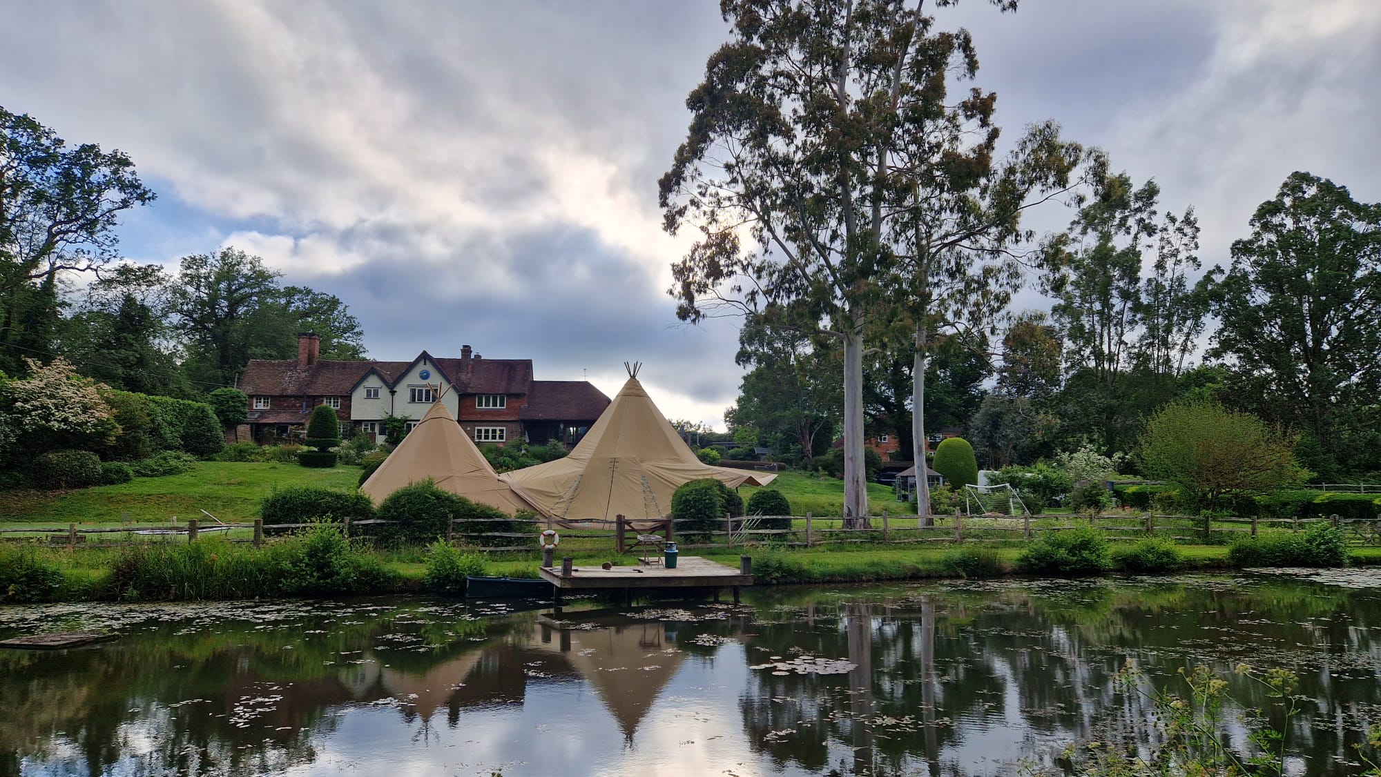 A tipi mirrored in the still water of an ornamental pond, with a period manor house and mature trees beyond
