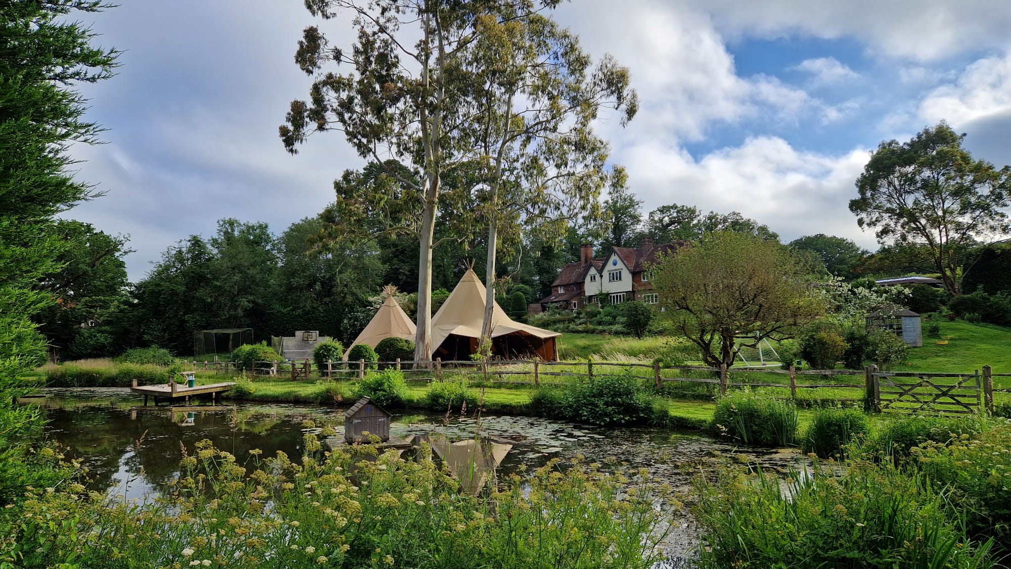 Twin linked tipis on the far bank of a wildlife pond with a tile-hung country house behind and lily pads in the foreground