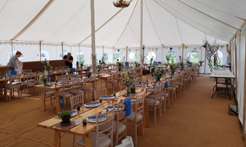 Interior of a traditional pole marquee during setup — unlined canvas visible, trestle tables with limewash chiavari chairs
