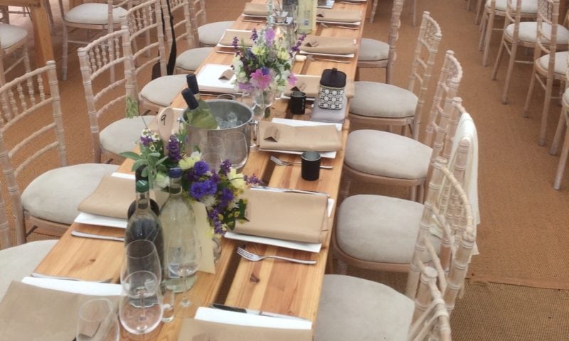 Bird's-eye close-up of a rustic trestle table with wildflower centrepieces, crystal glassware and limewash chiavari chairs